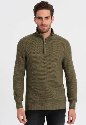 Suéter de punto verde oliva con cuello de media cremallera, superficie texturizada y puños y dobladillo acanalados. Llevado con jeans negros.