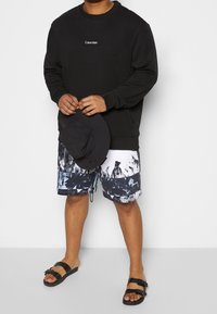 Sweatshirt noir à manches longues avec logo, shorts à motifs bleus et blancs, et chapeau noir à large bord ; personne portant des sandales noires.
