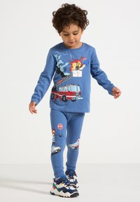Ein kleines Kind in blauen Schlafanzügen mit Grafiken von Feuerwehrautos, Flammen, Feuerwehrmann, Polizeiauto, Krankenwagen und Stoppschild, das drinnen läuft.