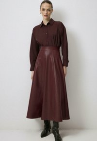 Sieviete stāv burgundijas krāsas pogātā krekla un atbilstošās augstajos jostā A formas ādas svārkos, kājās melnas augstpapēžu zābakus pret vienkāršu sienu.