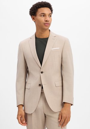 Homme portant une veste de costume beige et un pantalon sur une chemise vert foncé, regardant sur le côté sur un fond blanc uni.