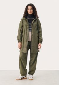 Jachetă verde măsliniu impermeabilă, cu manșete elastice, glugă și buzunare laterale, purtată împreună cu pantaloni elastici asortați și teniși albi.