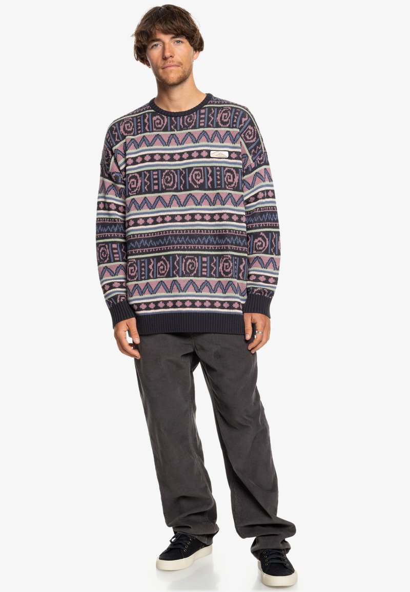 Multi-colored knit sweater featuring geometric patterns in pink, navy, and cream, with ribbed cuffs and hem; paired with gray corduroy pants.
