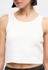 Γυναίκα που φοράει λευκή αμάνικη κοντή μπλούζα συνδυασμένη με μαύρο υφασμάτινο παντελόνι, εμφανιζόμενη από το πηγούνι μέχρι τη μέση σε απλό φόντο.