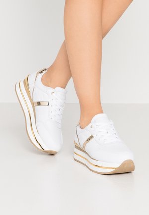 Weiße Plateau-Sneaker mit goldenen Akzenten, getragen an übereinandergeschlagenen Beinen vor einem schlichten hellen Hintergrund.