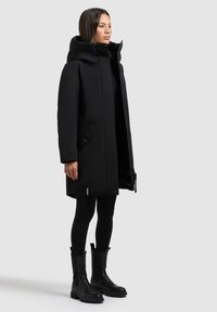 Sort isoleret parka med høj krave, lynlås fortil, sidelommer og en afslappet pasform, parret med sorte knælange støvler og leggings.