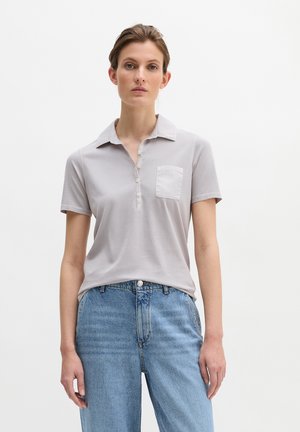 Frau trägt ein hellgraues Polo-Shirt mit kurzen Ärmeln und Brusttasche sowie hellblaue Jeans mit hoher Taille, steht vor einem schlichten weißen Hintergrund.