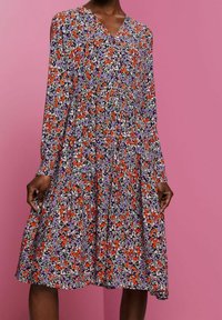 Pitkähihaista mekkoa, jossa on V-pääntie, purppuran-, oranssin- ja valkoisen kukkakuosisen mustan kankaan päällä, pukeutuneena henkilö seisoo vaaleanpunaisen taustan edessä.