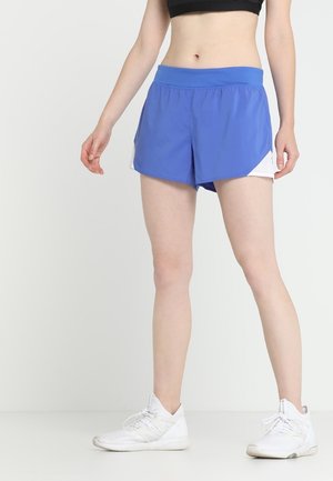 Kvinde iført blå sportsshorts og hvide sneakers, stående mod en ensartet lys baggrund.