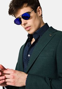 Zielona marynarka o teksturze, noszona na ciemnoniebieskiej koszuli z małym białym wzorem. Okrągłe niebieskie okulary przeciwsłoneczne, dłonie złożone, subtelne logo na marynarce.