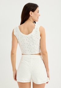 Top corto sin mangas de color blanco con bordes con volantes y un diseño de ojales bordados, combinado con pantalones cortos blancos de talle alto a juego con dobladillos en forma de ola.