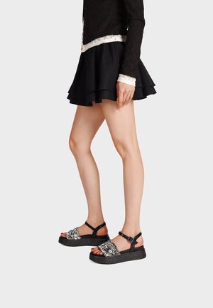 Donna che indossa una minigonna nera a strati, un maglione nero rifinito con pizzo e sandali con plateau neri con cinturini a motivo floreale bianco.