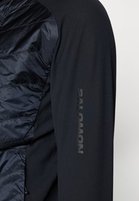 Primo piano della manica di una giacca nera con imbottitura trapuntata sulla spalla e logo "SALOMON" stampato verticalmente sul braccio.