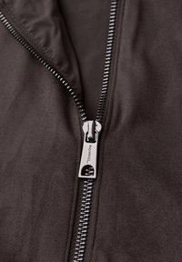 Gros plan sur une veste marron foncé partiellement zippée, avec un zipper en métal argenté gravé du logo "Street One".
