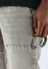 Pantalones cargo de mezclilla gris con múltiples bolsillos, herrajes plateados y una tela texturizada. Detalle de correa lateral visible cerca de la cintura.