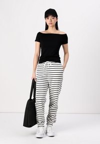 Črn off-shoulder top, bele črtaste trenirke, športni čevlji in črna kapa. Drži črno torbo, stoji na enobarvni podlagi.
