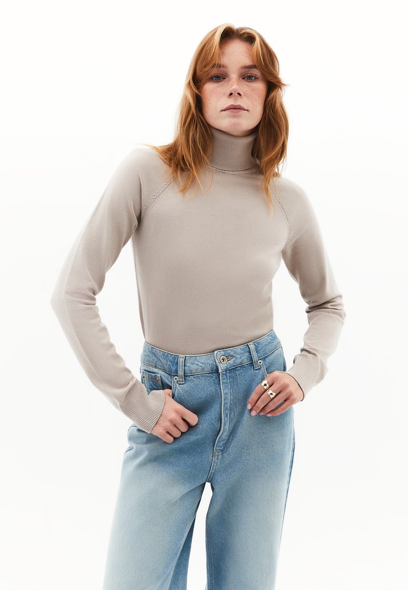 Beige coltrui met ribgebreide manchetten, gecombineerd met lichtblauwe high-waisted jeans. De trui is soepel, aansluitend en met lange mouwen.