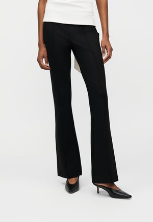 Femme portant un pantalon noir évasé, un haut blanc et des escarpins noirs à bout pointu avec talon kitten, debout devant un fond blanc uni.