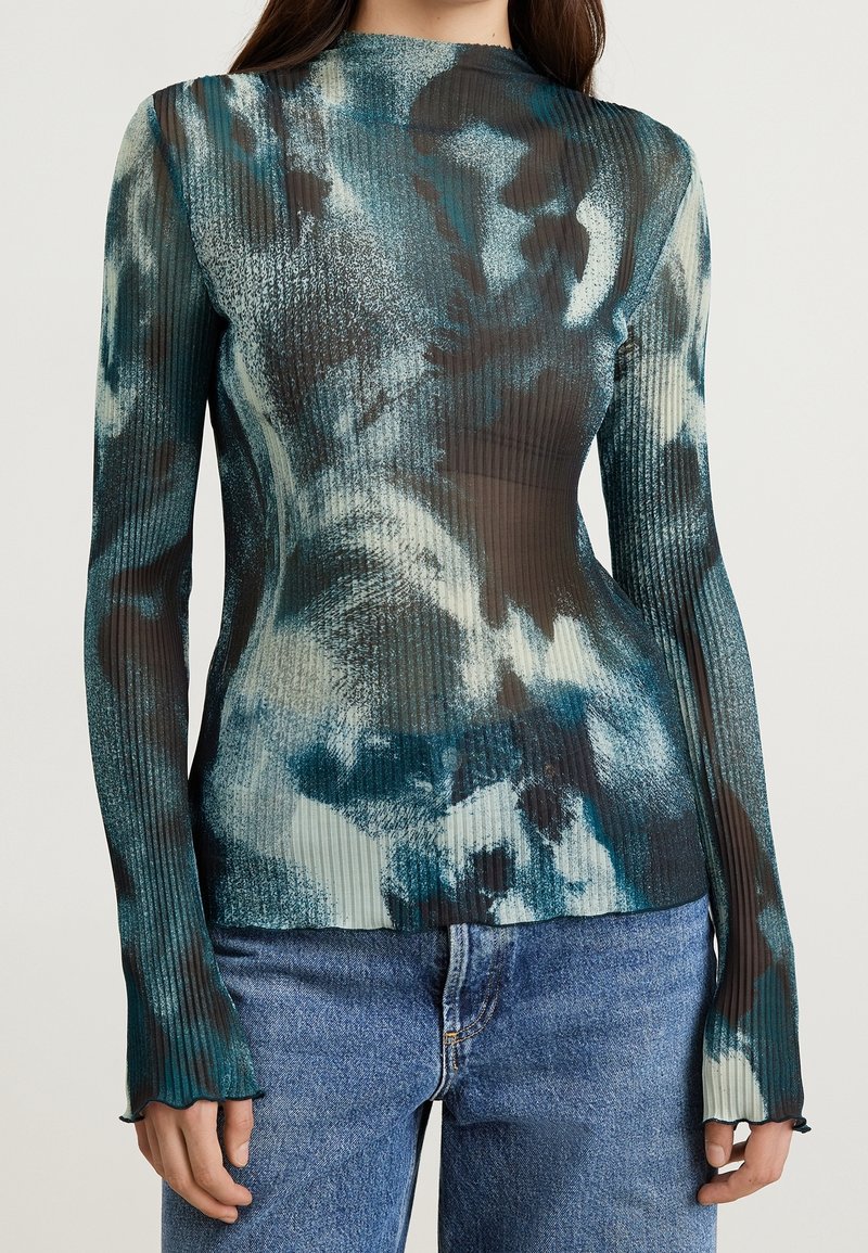 Long-sleeve top in ribbed fabric, featuring a blue and black abstract pattern, with a fitted silhouette and ruffled cuffs. Paired with blue jeans.