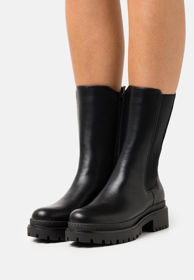 Bottes en cuir noires mi-mollet avec semelles en caoutchouc épaisses portées sur des jambes nues, présentées sur un fond clair uni.