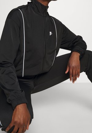 Persoon in een zwart Nike trainingsjack met witte biezen, zittend met één knie omhoog, handen rustend op de benen tegen een effen achtergrond.