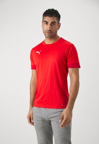 Camiseta atlética roja de manga corta, hecha de tela suave. Presenta un logo blanco de Puma en el pecho. Combinada con pantalones cortos grises.