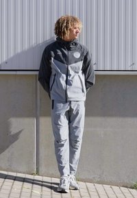 Grå og svart treningsdress med glidelåsjakke og bukser, med et lite logo og lommer, laget av lett materiale.