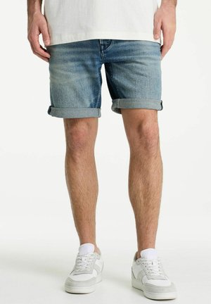 Man wearing rolled-up denim shorts, white sneakers, and white ankle socks, standing against a white background.