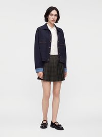 Jeune femme debout portant une veste bleu marine avec des poignets en denim, une chemise blanche boutonnée, une jupe plissée à carreaux et des chaussures Mary Jane noires.