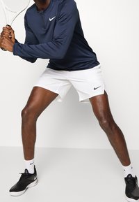 Tummansininen pitkähihainen urheilupaita pyöreällä kaula-aukolla ja valkoisella Nike-logolla; valkoiset shortsit; mustat urheilukengät; pitää tennismailaa käsissään.