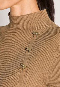Nahaufnahme einer Person, die einen gerippten, kamelbraunen Rollkragenpullover trägt, auf dessen Schulter sich drei kleine goldene Dekorationsbroschen in Form einer Schleife befinden.