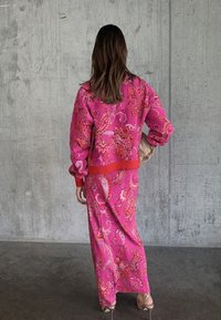 Γυναίκα με μακριά καστανά μαλλιά, ντυμένη με ροζ σακάκι με σχέδιο παϊέτας και μακριά φούστα που ταιριάζει, κρατώντας μια μπεζ μικρή τσάντα, στέκεται μπροστά σε σκυρόδετο τοίχο.