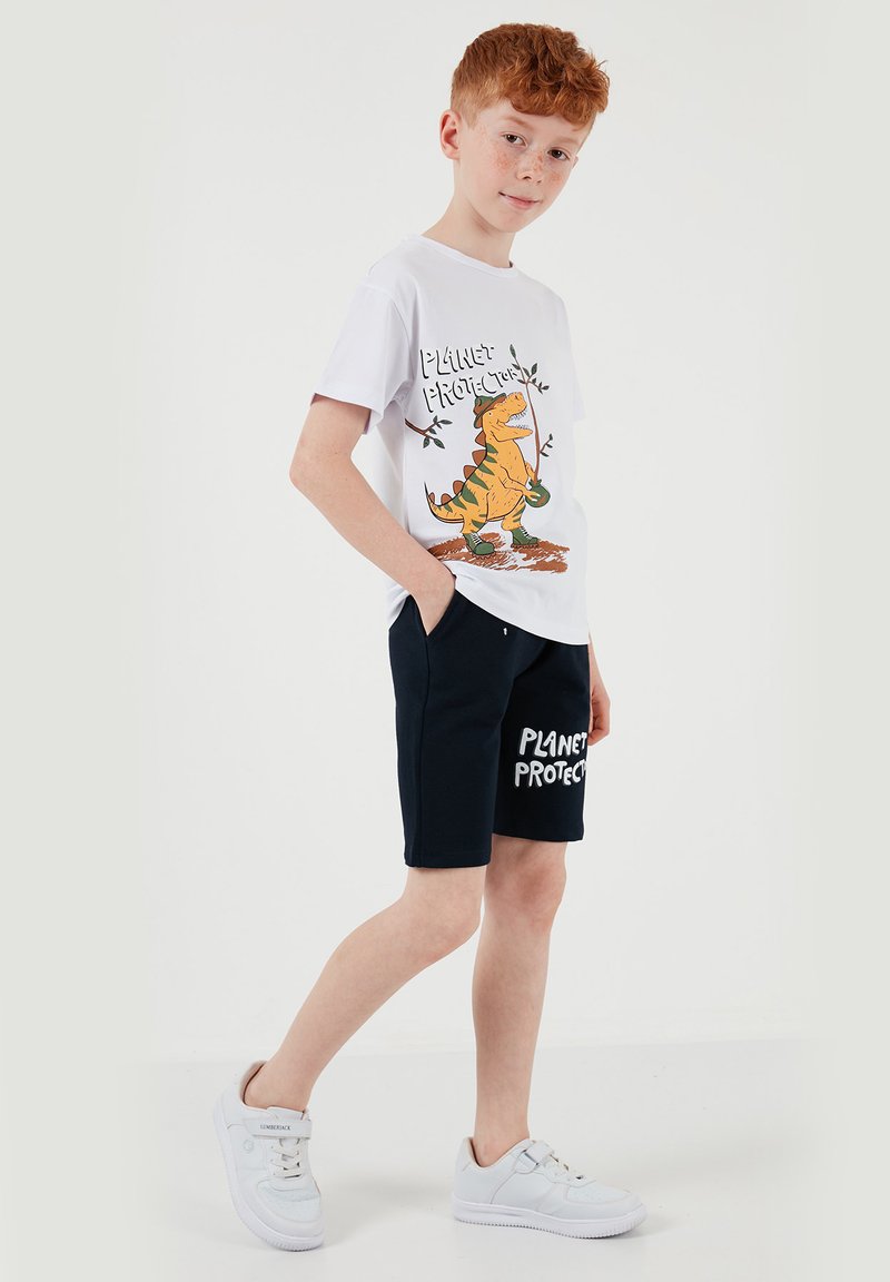 Garçon aux cheveux roux portant des baskets blanches, un short noir avec le texte « PLANET PROTECT » et un t-shirt blanc arborant un dinosaure et le design « PLANET PROTECTOR ».