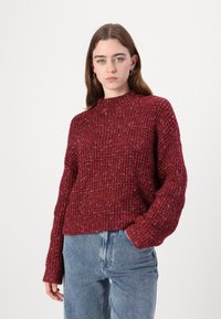 Πλεκτό κόκκινο πουλόβερ με ψηλή λαιμόκοψη, υφασμάτινο μοτίβο και μακριά μανίκια. Συνδυασμένο με ανοιχτό μπλε τζιν ψηλόμεσο.