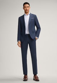 Granatowy dopasowany garnitur z marynarką zapinaną na jeden guzik, spodniami z płaskim przodem oraz białą koszulą. Brązowe skórzane buty typu double monk strap.