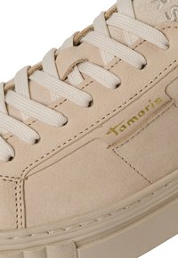 Baskets en daim beige avec lacets plats beiges, coutures visibles et nom de marque "Tamaris" en doré sur le panneau latéral.