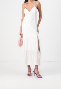 Mujer con vestido midi blanco sin mangas con abertura lateral, sosteniendo un bolso pequeño rosa y usando sandalias de tacón alto con tiras moradas.