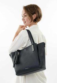 Zwarte leren totebag met een textuurafwerking, voorzien van dubbele handvatten, een ritsluiting en een decoratieve kwastdetail.