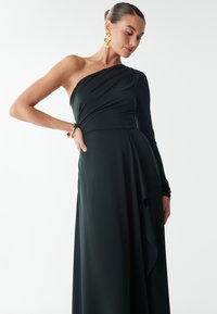 Één-schouder, vloer_length jurk in een donkergroene stof. Lange mouw aan één kant, vloeiende rok met drapering details. Gouden accessoires zichtbaar.