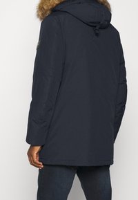 Námořnicky modrá parka s kapucí lemovanou kožešinou, dlouhými rukávy a bočními kapsami. Má texturovanou látku a vojenský odznak na rameni.