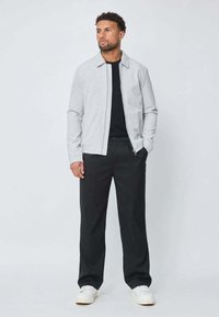 Homme se tenant avec les mains dans les poches, portant une veste zippée grise clair, un t-shirt noir, un pantalon noir et des baskets blanches sur fond uni.