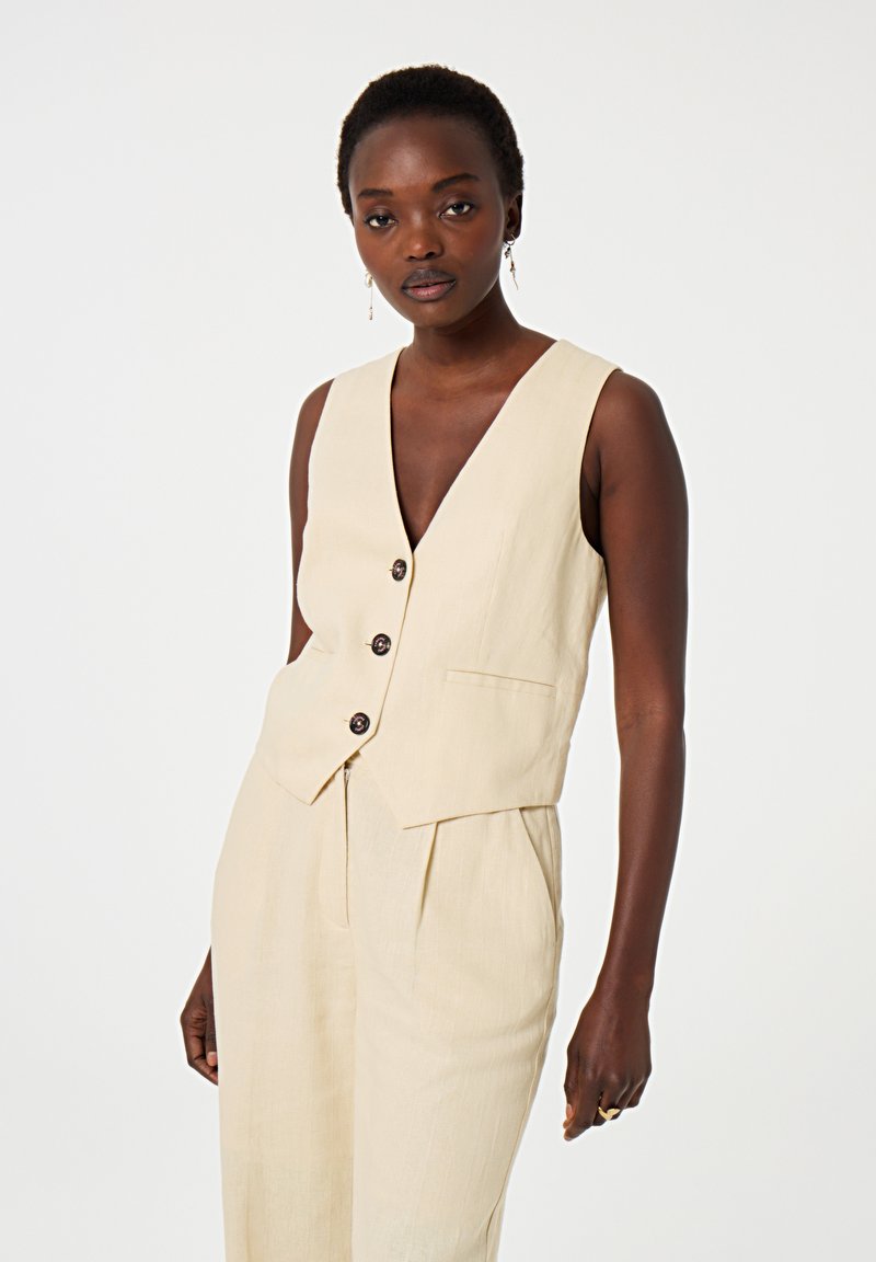 Beige Leinenweste mit tiefem V-Ausschnitt, fünf Knöpfen, Seitentaschen und taillierter Passform, kombiniert mit passenden, lockeren Hosen.