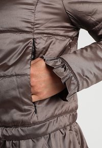 Brązowa kurtka puchowa o błyszczącej powierzchni, z falbanastym mankietem i skośną boczną kieszenią z czarnym suwakiem.