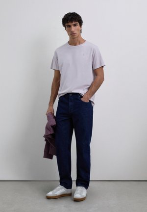 Jeune homme portant un t-shirt violet clair, un jean bleu foncé, des baskets blanches, tenant un sweat à capuche violet replié, se tenant devant un mur blanc uni.