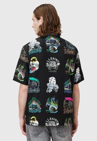 Camisa de manga corta negra con patrones gráficos coloridos, incluyendo calaveras, dragones y palmeras, con un corte relajado y cierre de botones.