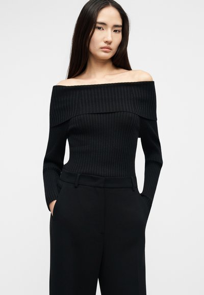 Vrouw met lang donker haar, gekleed in een zwarte, off-shoulder geribbelde trui en zwarte, hooggetailleerde broek met handen in de zakken.