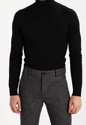 Homme portant un pull à col roulé noir ajusté et un pantalon de costume gris texturé, debout avec les bras relâchés sur les côtés.