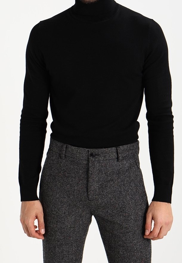 Homme portant un pull à col roulé noir ajusté et un pantalon de costume gris texturé, debout avec les bras relâchés sur les côtés.