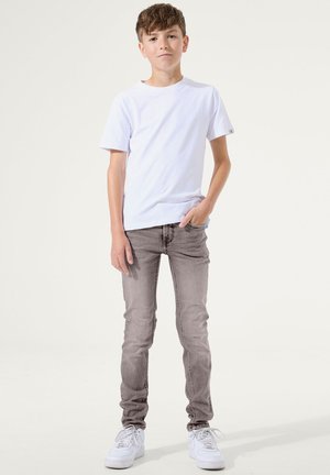 Junge trägt ein weißes T-Shirt, graue Jeans und weiße Sneaker, steht mit einer Hand in der Tasche vor einem schlichten hellen Hintergrund.