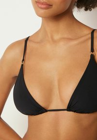 Mujer con un top de bikini negro en forma de triángulo con tiras delgadas y detalles de anillos dorados, mostrando el cuello, los hombros y la parte superior del pecho.